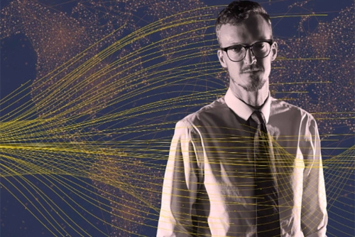 Dark image of a man dressed in a shirt and tie stands in front of a map of the world with lines representing data flows over images. 