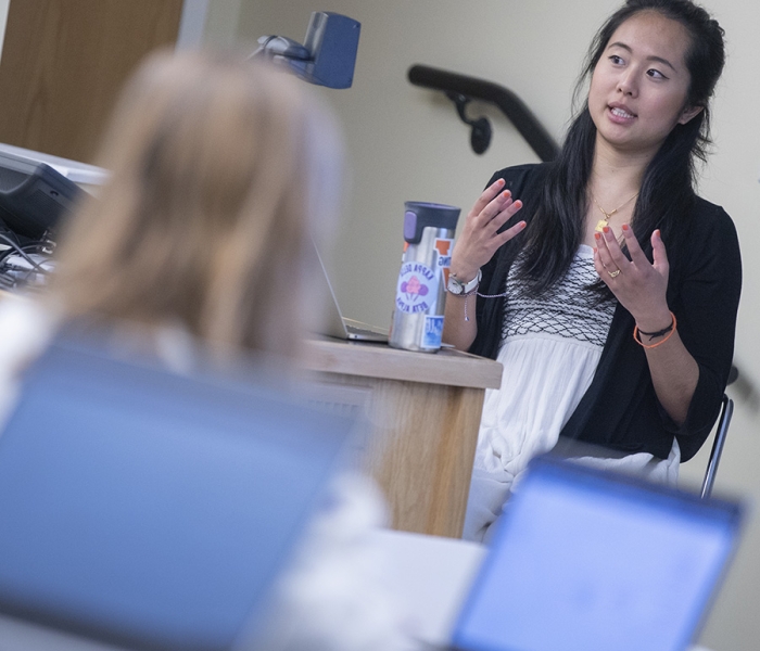 Female professor teaching in front of classroom of students at laptops